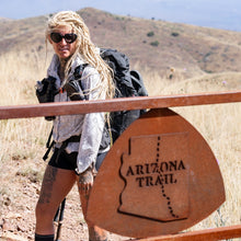Load image into Gallery viewer, A woman with long blonde dreadlocks and sunglasses hikes behind a rusted metal gate marked Arizona Trail, wearing a gray patterned shirt, black shorts, and a black backpack in a dry grassy mountain setting.
