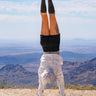A person performing a handstand outdoors on a mountain ridge under a blue sky with clouds, wearing black leggings, a white long-sleeve top, and gray sneakers.