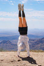 Load image into Gallery viewer, A person performing a handstand outdoors on a mountain ridge under a blue sky with clouds, wearing black leggings, a white long-sleeve top, and gray sneakers.
